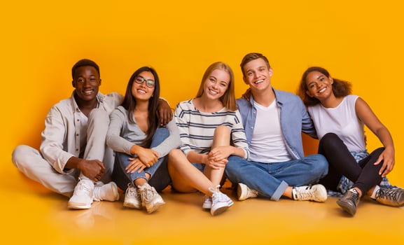 Multiracial joyful students sitting on floor, embracing and having fun over yellow studio background, students lifestyle concept, panorama photo