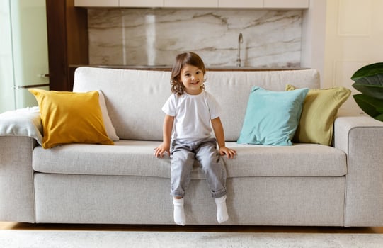 Cute preschool little girl sitting on couch at home, smiling at camera with innocence and joy, adorable female child embodying childhood happiness in comfortable living room interior, copy space photo