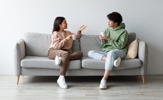 Young Asian boyfriend and girlfriend drinking coffee and chatting while sitting on sofa at home. Affectionate millennial couple enjoying hot drink, having conversation, spending time together photo