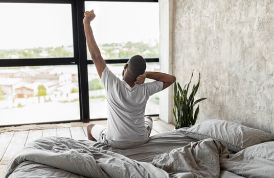 Good Early Morning Concept. Rear Back View Of African American Male Waking Up Sitting On Bed In Bedroom At Home. Millennial Man Stretching Back And Arms, Looking At Window, Free Copy Space photo