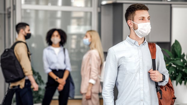 Social distance and work in office after covid-19 quarantine. Focus on millennial man with protective mask and backpack, looks to the side in hallway, and workers are blurred, free space photo