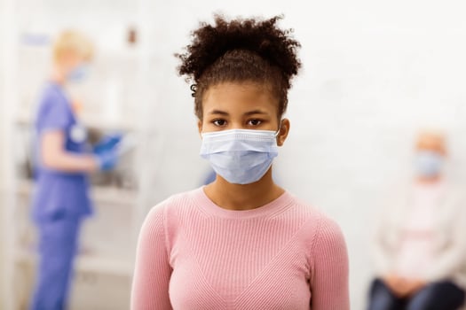 Portrait Of Black Teen Girl Patient Infected With Coronavirus Posing Wearing Medical Mask Standing In Hospital Clinic Room, Looking At Camera. Covid-19, Corona Virus Disease Concept photo