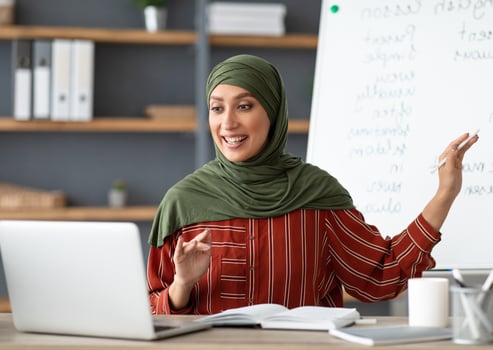 Online Education Concept. Portrait of smiling female arabic teacher in hijab pointing at whiteboard, explaining English grammar rules to students. Excited woman looking at laptop screen, talking photo