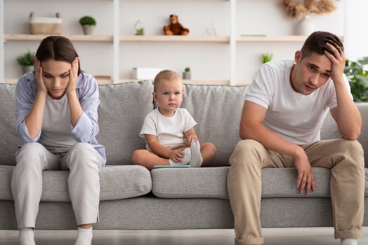 Family Conflicts. Cute infant baby sitting between offended parents after quarrel at home, young spouses with child suffering marital crisis, feeling depressed and frustrated, free space photo