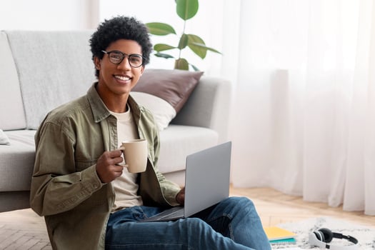 Positive black teen guy sitting on floor with laptop and cup of coffee, studying online from home, copy space. African American teenage student enjoying hot beverage during educational webinar photo