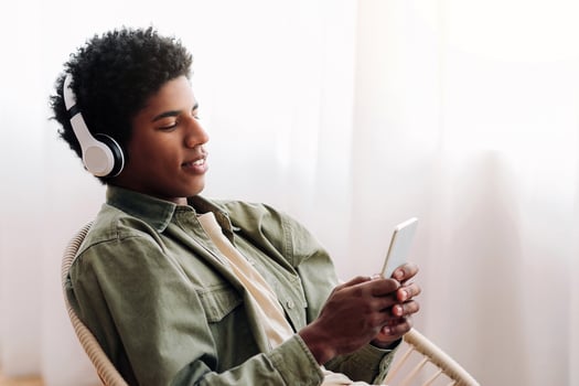 Attractive black teen in headset choosing music playlist on cellphone at home. Funky African American guy downloading his favorite songs on mobile device, listening to audiobook or radio photo