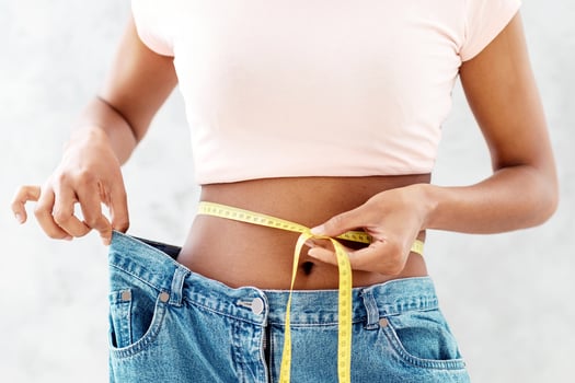 Closeup of black woman in old big jeans measuring her waist with tape, showing results of slimming diet or liposuction, grey background. Young lady promoting healthy nutrition for weight loss photo