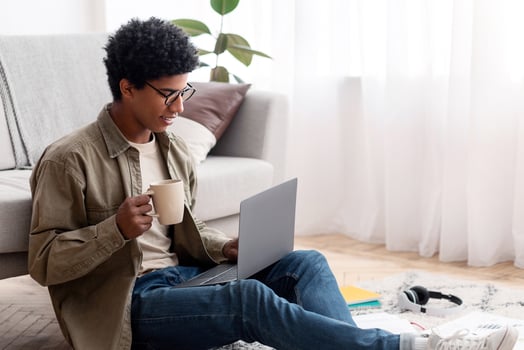 Attractive black teenager using laptop and drinking coffee during his online studies at home, copy space. African American teen guy making home assignment, writing coursework, taking part in webinar photo