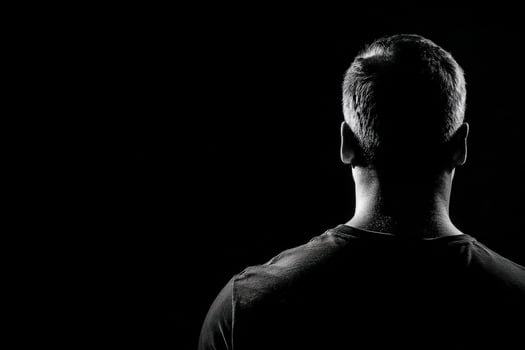 A man stands facing away from the viewer, showcasing a silhouette against a stark black background. photo