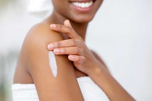 Beautiful skin and spa day at home during covid-19 quarantine. Young smiling african american female in white towel smears body with moisturizer means in bedroom interior, cropped, close up photo