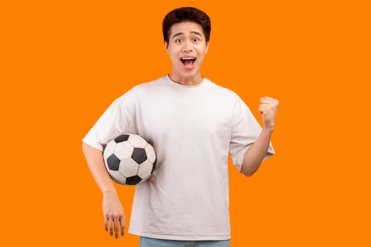 Go On. Portrait of excited young asian man making winner gesture, watching football game, standing and holding soccer ball, isolated over yellow studio background, banner. Guy cheering and screaming photo