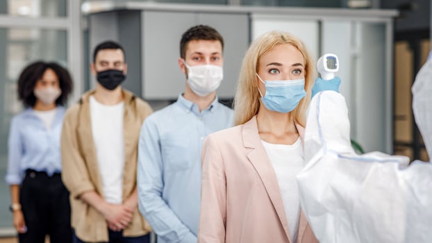Temperature control test at work and social distancing. Man in protective suit checks multinational workers with non-contact thermometer in morning at entering in office, empty space photo