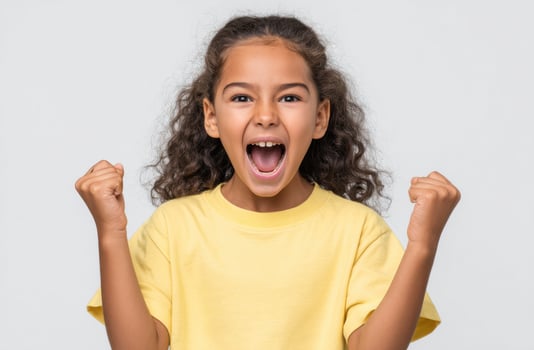 A jubilant young girl with curly hair, wearing a yellow shirt, expresses unrestrained joy and excitement with raised fists. Her be photo