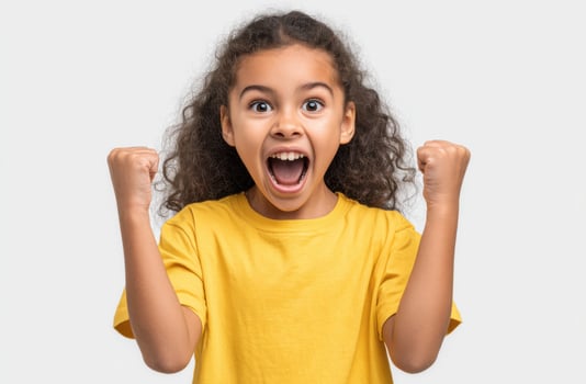 A jubilant young girl with curly hair, wearing a yellow shirt, expresses pure elation with open mouth and raised fists. This portr photo