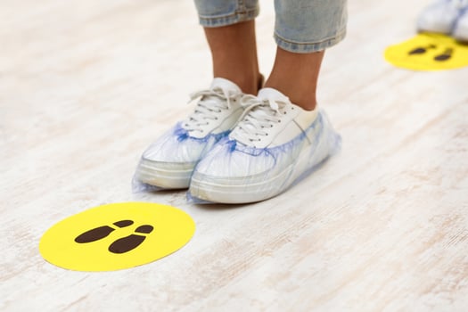 Keep Safe Distance. Teen Girl's Feet With Shoes Covers Standing Near Social Distancing Warning Sign On Floor In Queue Line Indoors. Covid-19, Coronavirus Protection Background. Cropped photo