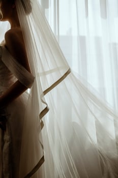 Silhouette of a blonde bride with a lush veil, in a long white dress near a window with a bouquet of lilies. Minimalistic interior, hotel or house. Bride in a long dress photo
