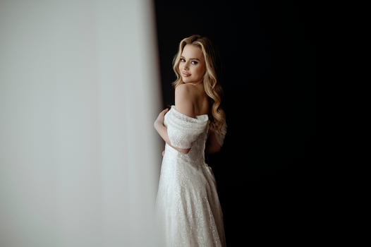 Elegant bridal gown showcased in a minimalist setting with a model in a chic pose during an indoor shoot. image of a blonde bride against a dark interior. smiles happily and looks at the camera photo