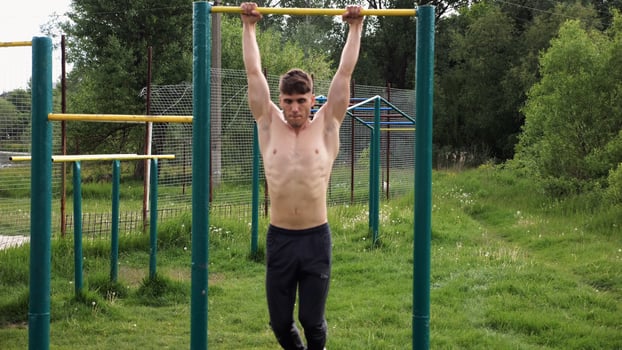 Muscula athlete doing pull ups on horizontal bar at sports ground. Young shirtless sportsman exercising at nature. Strong sporty man training outdoor. Concept of healthy and active lifestyle. Slow mo photo