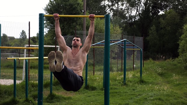 Athletic man train abdominal muscles raising legs on horizontal bar at sports ground. Young sportsman exercising on pull-up bar outdoor. Sporty athlete working out at nature. Healthy lifestyle photo