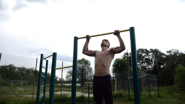 Muscular athlete doin pull ups on horizontal bar at sports ground. Young shirtless sportsman exercising at nature. Strong sporty man training outdoor. Concept of healthy and active lifestyle. Slow mo photo