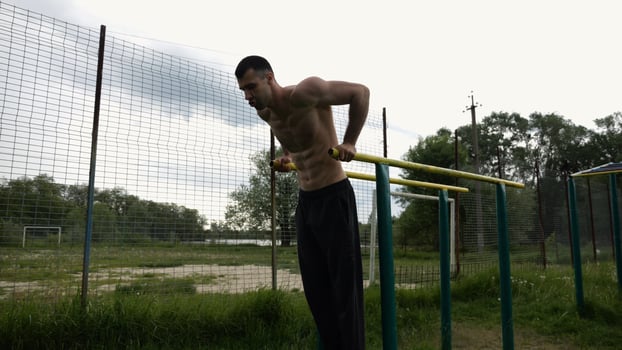 Young sportsman doin push ups on parallel bars at nature. Strong sporty man exercising at sports ground. Male shirtless athlete training hard outdoor. Concept of active sportive lifestyle. Slow mo photo