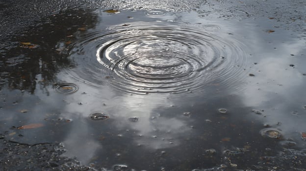 Raindrops falling create ripples in a puddle reflecting the sky adding texture to the asphalt surface photo