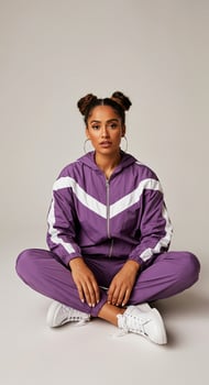 Young woman in purple athletic tracksuit sits cross-legged on the floor, showcasing a relaxed pose, with a neutral background emphasizing her casual style and confidence photo