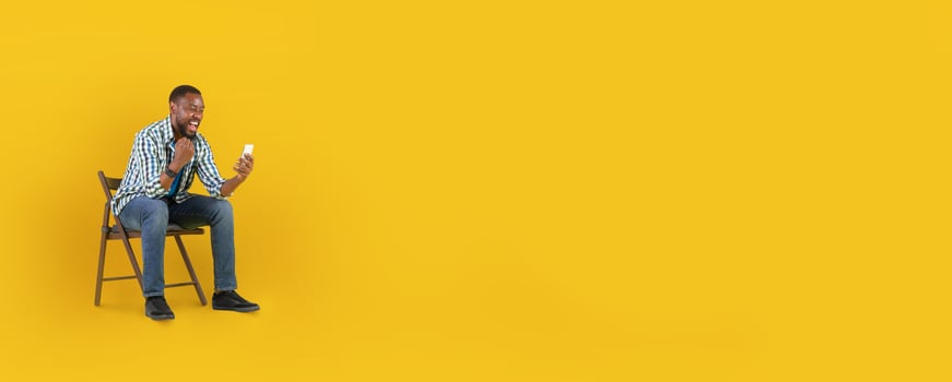 African American man is sitting on a wooden chair against a bright yellow background, laughing joyfully while looking at his smartphone. His expression shows delight and engagement. photo