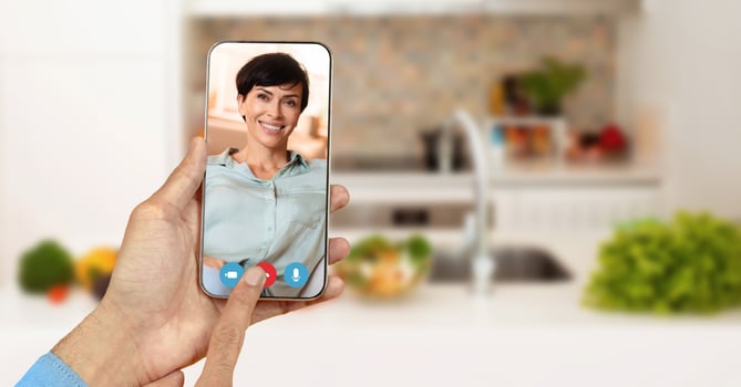 A person holds a smartphone with smiling face during a call in a cozy kitchen. Fresh ingredients and a homey atmosphere create a casual setting for communication and connection. photo