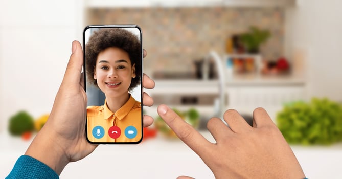 A hand holds a smartphone displaying a smiling person on the screen during a call. The background shows a bright kitchen with fresh vegetables, creating a lively atmosphere. photo