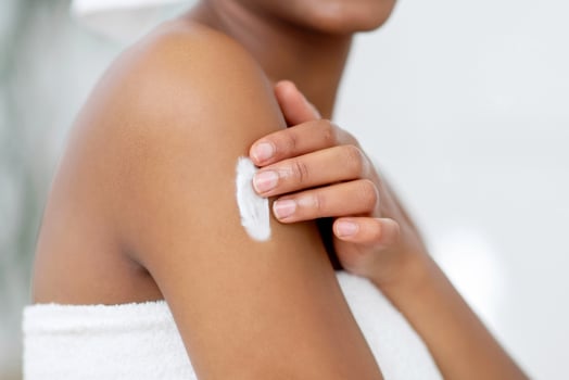 Moisturizing skin in morning after shower at home. Happy millennial african american female wrapped in white towel takes care of body in modern bedroom interior, cropped, close up, free space photo
