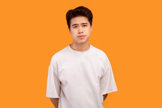 Male Model. Portrait of asian guy wearing blank white t-shirt for design print mockup, standing isolated over orange studio background. Calm casual teenager posing and looking at camera photo