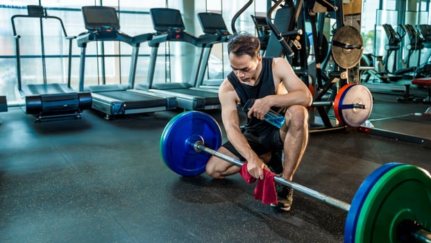Asian man wiping barbell with red towel in gym after workout, practicing cleanliness and hygiene while holding a cleaner solution in fitness environment photo