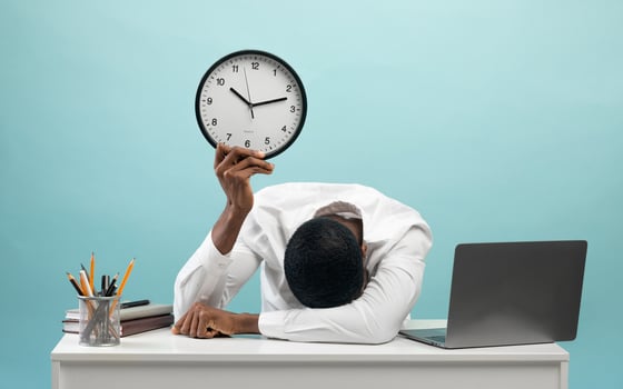 Tired african american businessman sitting at desk with pc laptop, holding clock and putting head on hand, feeling exhausted over blue studio background photo