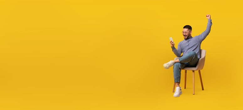 Asian man sitting on a chair beams with joy as he raises his fist in celebration while looking at his smartphone against a vibrant yellow backdrop. photo