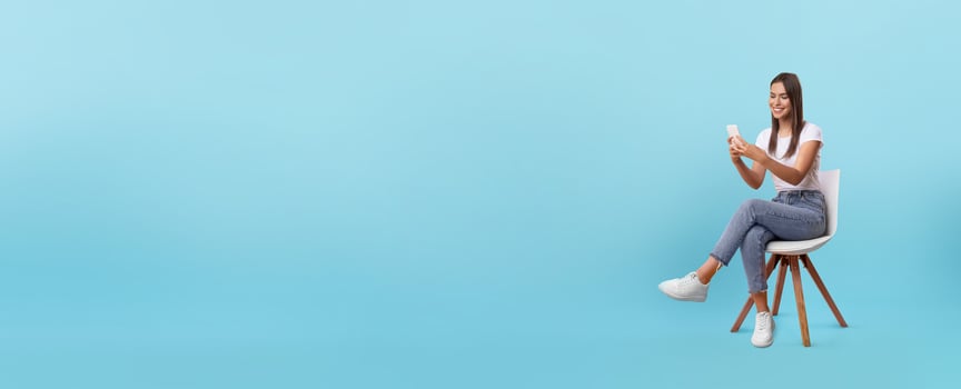 A young woman sits comfortably on a chair, absorbed in her smartphone. She wears casual clothing and smiles at the screen, with a simple blue background enhancing the scene. photo