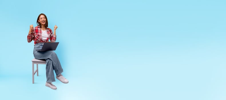 Asian woman sits on chair, smiling and raising her hands in triumph as she uses a laptop. The bright blue background adds a cheerful vibe to her joyful moment. It captures a sense of accomplishment. photo