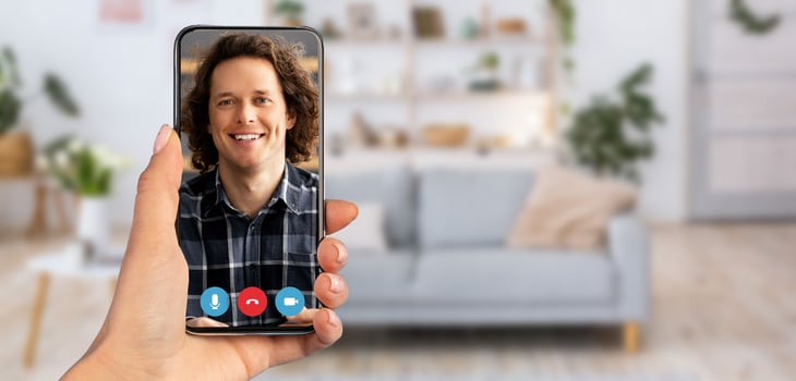 A hand holds a smartphone displaying a man smiling during a call. The setting features a cozy home interior with a modern couch and plants in the background, creating a relaxed atmosphere. photo
