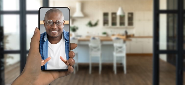 A person holds a smartphone displaying a smiling individual during a call. The setting features a bright kitchen with wooden floors and a modern, inviting atmosphere. photo