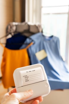 Hand holding mini thermal printer with clothing decluttering checklist in bright minimalist room with organized garments on hangers in background concept of smart home wardrobe planning and sorting photo
