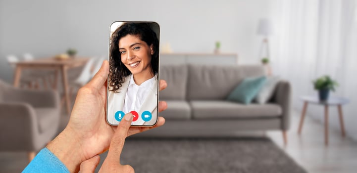 A person engages in a call from home, displaying a screen with smiling face. The setting includes a cozy living room with a sofa and soft furnishings, creating a friendly atmosphere. photo