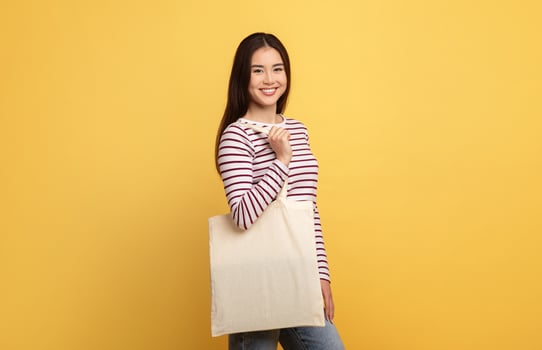 A young Asian woman stands against a cheerful yellow backdrop, smiling while holding a simple tote bag. She wears a stylish striped long-sleeve shirt and casual jeans, radiating positivity. photo