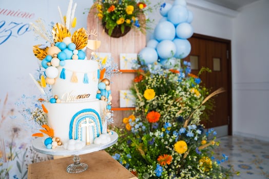 A multi tiered cake with blue, orange, and gold decorations, surrounded by floral arrangements and balloons, set in an indoor festive setting. photo