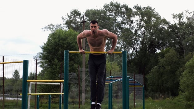 Hardy shirtless sportsman doing push up on horizontal bar at nature. Strong and muscular guy training at sports ground. Young athletic guy exercising outdoor. Concept of active and healthy lifestyle photo