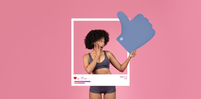 A young woman in a cropped top and shorts expresses surprise while pointing at a large thumbs-up symbol. The vibrant pink backdrop enhances her excitement, showcasing social media interaction. photo