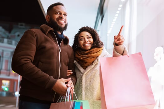 Sales And Discount. Cheerful curly black woman pointing finger at mall window, indicating showcase, walking with her man in the evening near shopping center, holding colorful bags with purchases photo