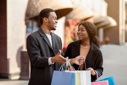 Love, holiday sales, shop, retail, consumer. Cheerful millennial african american man and lady with many coloured shopping packages, guy shows things in bag after shopping in city mall, free space photo