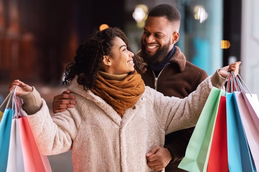 Couple In Love. Cheerful handsome african american guy hugging his girlfriend or wife who holding colorful shopping bags and looking at him. Happy black lovers having fun outdoors and laughing photo