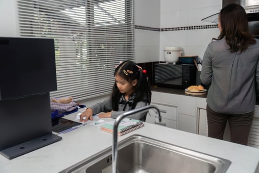 Parent-Child Bonding. Young girl studying at home while mother prepares breakfast. photo