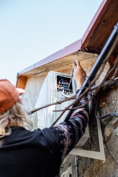 A man mounts an outdoor air conditioner unit on the wall of a stone farmhouse, installing electrical wiring and connecting a signal cable. photo